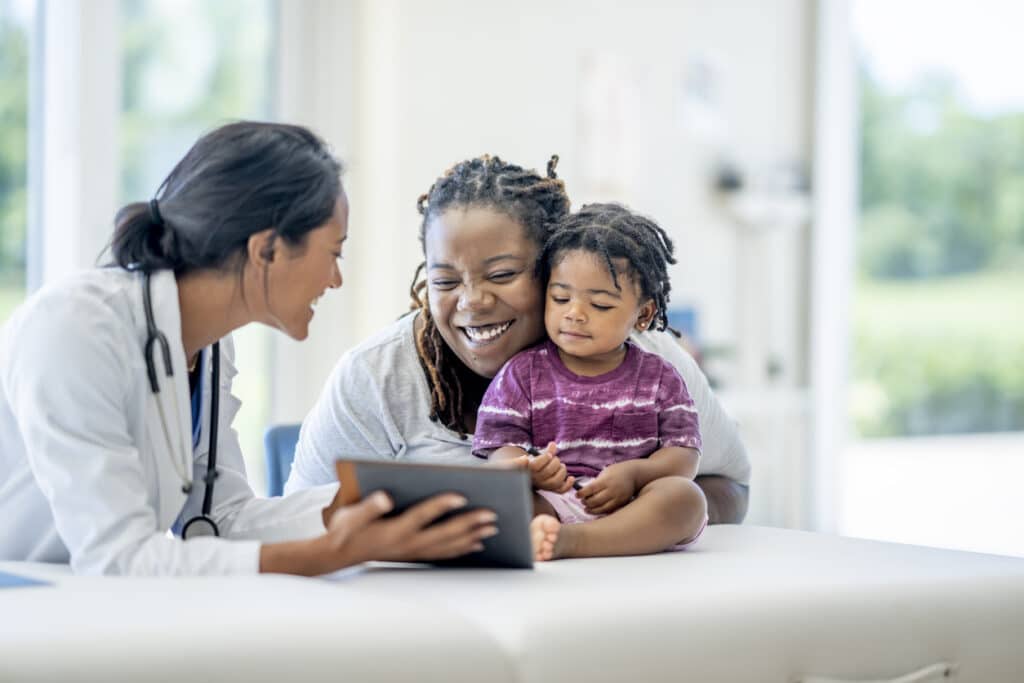 A young Mother and her son visit the doctor together.  The baby boy is seated on the exam table with his mother seated in front of him and making sure he does not fall.  Their female doctor of Middle Eastern decent is seated in front of them and smiling as they talk.